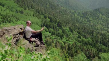 Dağlık bir bölgede kayaların üzerinde oturan ve telefonuyla güzel manzaraları fotoğraflayan genç bir adamın yan görüntüsü. Seyahat ve turizm