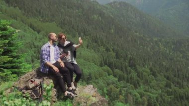 Genç bir adam ve bir kız dağlık bir bölgede bir kayanın üzerinde oturuyorlar ve telefonlarından selfie çekiyorlar. Seyahat