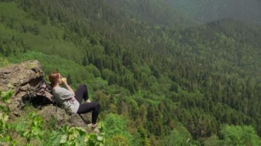 Genç bir kadın bir kayanın üzerinde oturuyor dağlık bir alanda elma yiyor ve manzaranın tadını çıkarıyor. Seyahat ve turizm