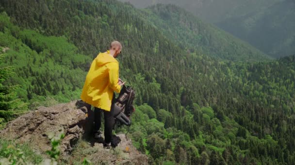 Genç bir adam çerçeveden sarı bir yağmurlukla ve sırtında sırt çantasıyla dağlık bir alanda çıkar. Seyahat ve turizm — Stok video