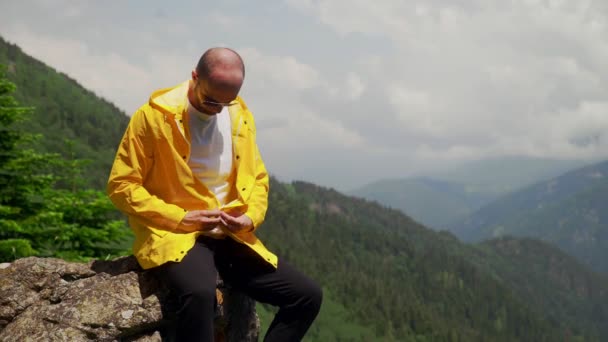 Um jovem de óculos escuros, sentado em uma rocha e abotoando uma capa amarela. Turismo e viagens — Vídeo de Stock