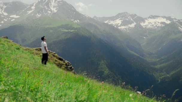 Siyah gözlüklü genç bir adam yamaçta duruyor ve dağlık bölgenin muhteşem manzarasının tadını çıkarıyor. Turizm ve seyahat — Stok video