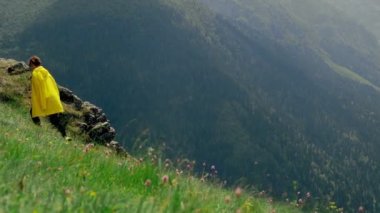 Sarı yağmurluk giymiş genç bir kadın yamaca tırmanıyor ve dağlık alanların muhteşem manzarasının tadını çıkarıyor. Turizm ve seyahat