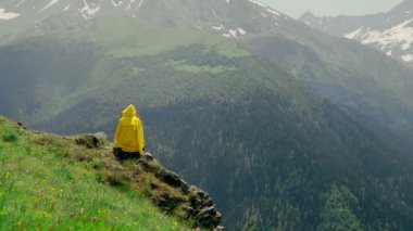 Sarı yağmurluklu genç bir kadın bir uçurumda oturuyor ve dağlık bölgelerin muhteşem manzarasının tadını çıkarıyor. Turizm ve seyahat