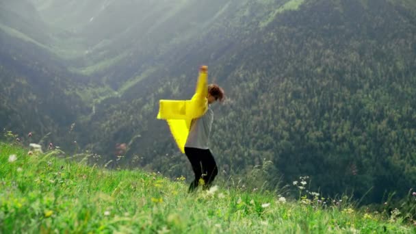 Sarı yağmurluk giymiş genç bir kadın kendi etrafında daireler çiziyor ve dağlık bölgelerin muhteşem manzarasının tadını çıkarıyor. Turizm ve seyahat — Stok video