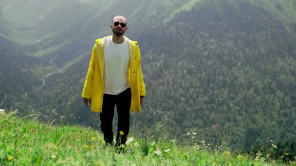 Sarı yağmurluk giymiş genç bir adam yamaca tırmanıyor ve dağlık alanların muhteşem manzarasının tadını çıkarıyor. Turizm ve seyahat — Stok video