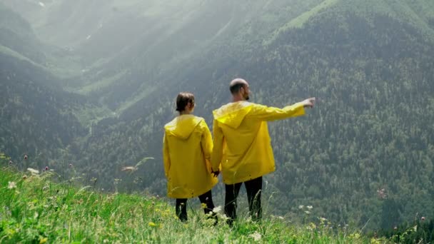 Un giovane uomo e una donna in impermeabili gialli sono in piedi sul pendio, tenendosi per mano e godendo il paesaggio della zona montuosa. Viaggi e turismo — Video Stock