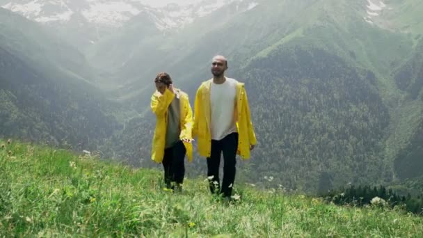 Un joven y una mujer con impermeables amarillos están subiendo por la ladera, tomados de la mano y disfrutando del paisaje de la zona montañosa. Viajes y turismo — Vídeo de stock