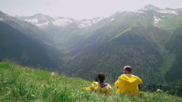 Egy fiatal férfi és egy nő sárga esőkabátban fekszenek a lejtőn, élvezve a hegyvidéki tájat. Utazás és turizmus — Stock videók