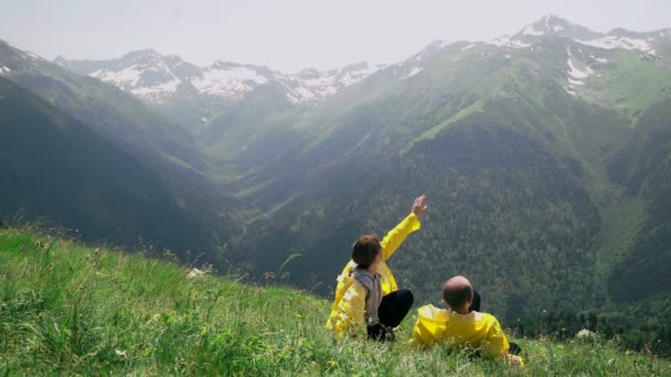 Egy fiatal férfi és egy nő sárga esőkabátban fekszenek a lejtőn, élvezve a hegyvidéki tájat. Utazás és turizmus — Stock videók