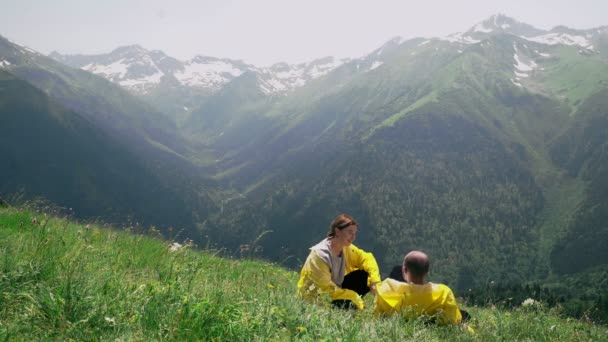 Um jovem e uma mulher de capas de chuva amarelas estão deitados na encosta, desfrutando da paisagem da área montanhosa. Viagens e turismo — Vídeo de Stock