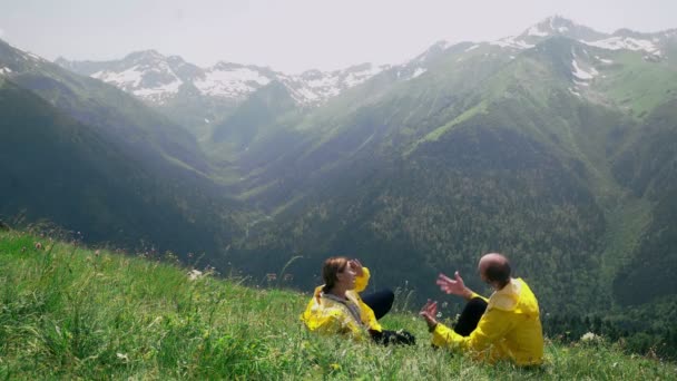 Sarı yağmurluk giymiş genç bir adam ve bir kadın yamaçta uzanmış dağlık bölgenin manzarasının tadını çıkarıyorlar. Seyahat ve turizm — Stok video