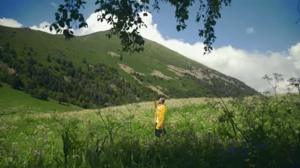 一个身穿黄色雨衣的年轻人站在一片草地上，欣赏着山区的风景。旅行和旅游 — 图库视频影像