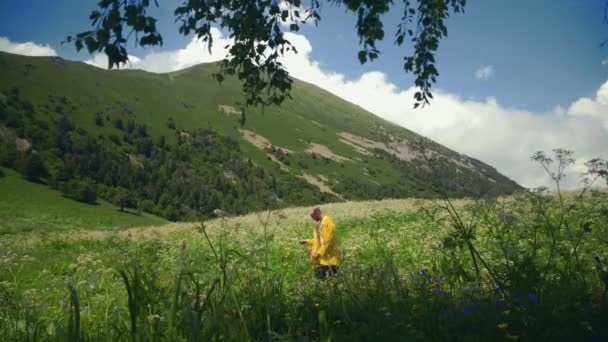 Mladý muž ve žlutém plášti trhá květiny na louce a užívá si scenérie v hornaté oblasti. Cestování a cestovní ruch — Stock video