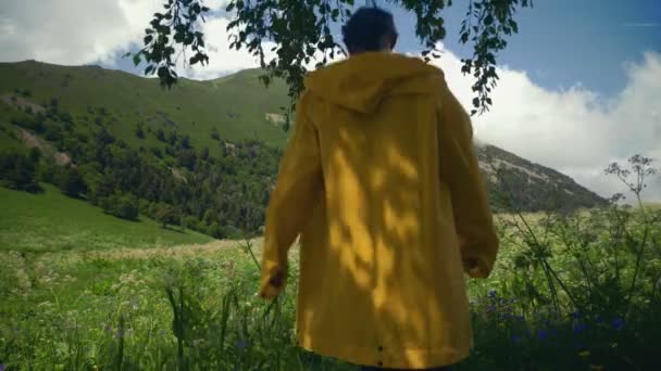 Sarı yağmurluk giymiş genç bir kadın çayırda yürüyor ve dağlık bir bölgedeki manzaranın tadını çıkarıyor. Seyahat ve turizm — Stok video