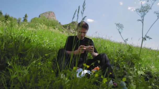 A young man is sitting on a slope and playing a game on his phone in a mountainous area. Tourism and travel — Stock Video