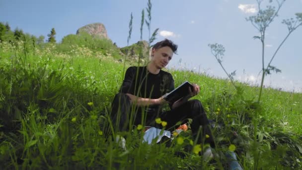 若い男が坂に座って本を読んで、山岳地帯の壮大な景色を楽しんでいます。旅行・観光 — ストック動画