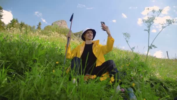Ein junger Mann in gelbem Regenmantel sitzt an einem Hang und kommuniziert per Video auf seinem Handy in einer bergigen Gegend. Tourismus und Reisen — Stockvideo