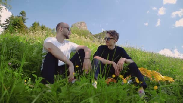 Due giovani con occhiali scuri sono seduti su un pendio e chattano in una zona montuosa. Turismo e viaggi — Video Stock