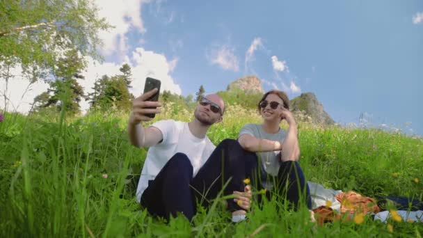 Een jonge man en een meisje zitten op een helling in een bergachtig gebied en nemen selfies op hun telefoon. Reizen — Stockvideo
