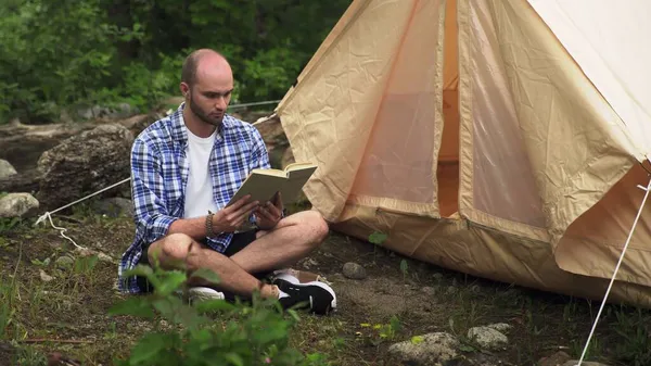 若者がテントの近くの地面に座り、森の中で本を読んでいる。観光とレジャー — ストック写真