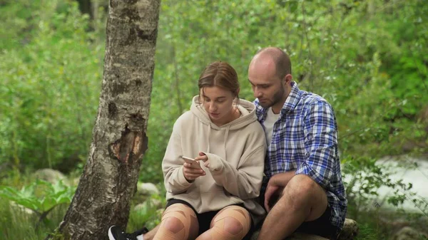 Un hombre y una mujer están sentados en una roca. Una mujer le muestra a un hombre algo en su teléfono. Un chico y una chica tienen una cita. En el bosque al aire libre — Foto de Stock