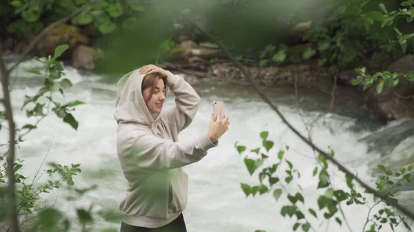 ヨーロッパ人の少女が山の川のほとりの森の中に立ち、自分の携帯電話で自撮りをする。路上で。観光 — ストック写真