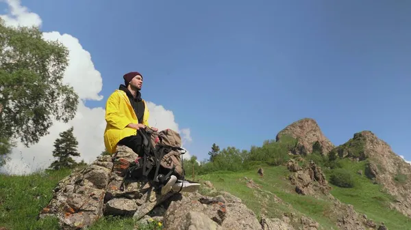 黄色のレインコートを着た男性観光客は、背中に大きなバックパックを持って岩の上に座って自然を楽しんでいます。観光. — ストック写真