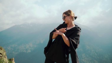 Güneş gözlüklü ve siyah kapüşonlu mutlu bir genç kız dağların arkasında dans ediyor.