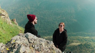 Genç bir adam ve bir kız dağlık bir bölgede bir kayanın üzerinde iletişim kuruyor ve güzel manzaralara bakıyor. Turizm