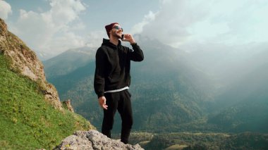 Dağlarda duran genç bir gezgin, telefonuyla internetten insanlarla sohbet ediyor ve arka plandaki nefes kesici manzarayı paylaşıyor..
