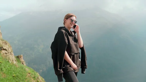 A happy young girl in sunglasses and a black hoodie is talking on the phone against the background of mountains and smiling. — Stock Photo, Image