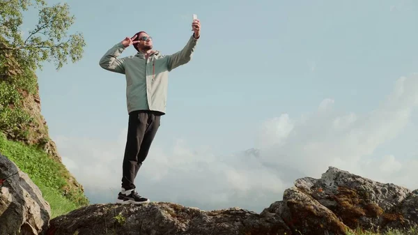 帽子とサングラスの若い男が岩の上に立って、自分の携帯電話で自撮りしている。旅行 — ストック写真