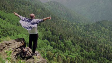 Genç bir adam dağlık bir bölgede bir kayanın üzerinde duruyor ve uzaklığa bakıyor. Seyahat ve turizm
