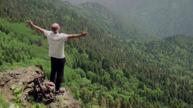 Genç bir adam dağlık bir bölgede bir kayanın üzerinde duruyor ve uzaklığa bakıyor. Seyahat ve turizm