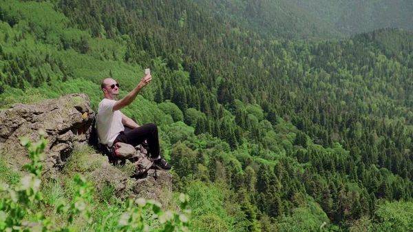 山間部の岩の上に座っている若い男のサイドビューと彼の携帯電話で美しい風景を撮影。旅行・観光 — ストック写真