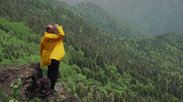 黄色のレインコートを着た若い男が山岳地帯の岩の上に立って、遠くを見ている。旅行・観光 — ストック写真