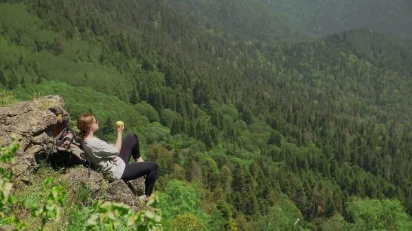 Eine junge Frau sitzt auf einem Felsen, isst einen Apfel in einer bergigen Gegend und genießt die Aussicht. Reisen und Tourismus — Stockfoto