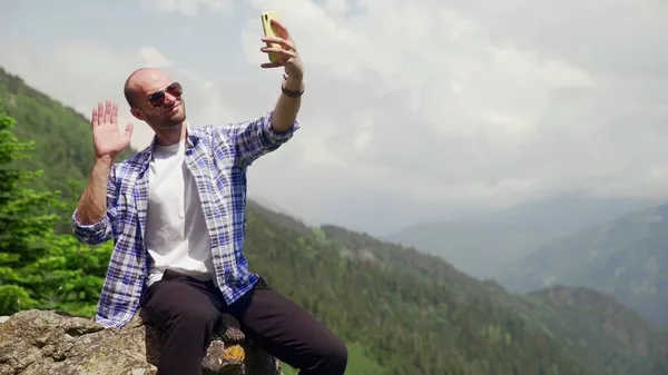 Um jovem de óculos de sol sentado sobre pedras e tirando uma selfie em seu telefone. Turismo e viagens — Fotografia de Stock