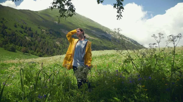 黄色のレインコートを着た若い女性が花束を持って牧草地に立っており、山間部の景色を楽しんでいます。旅行・観光 — ストック写真