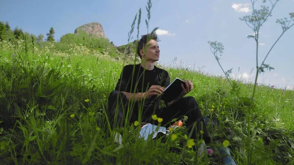 Um jovem está sentado em uma encosta, lendo um livro e apreciando a magnífica paisagem das regiões montanhosas. Turismo e viagens — Fotografia de Stock