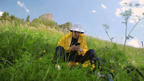 黄色のレインコートを着た若い男が斜面に座って、山間部の電話でテキストメッセージを送っています。旅行・観光 — ストック写真