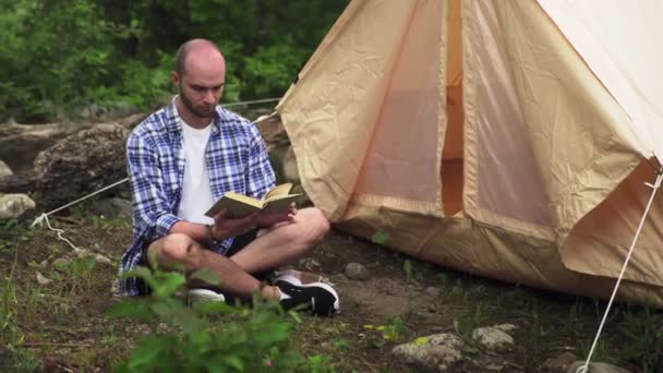 En ung man sitter på marken nära ett tält och läser en bok i naturen i skogen. Turism och fritid — Stockvideo