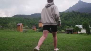 Dişi bir turist dağların ve ahşap evlerin arasında çimenlerde yürüyor. Doğa ve dağlar