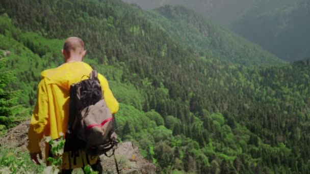 Mladý muž ve žlutém plášti a s batohem na zádech se přiblíží k útesu v hornaté oblasti a podívá se na nádhernou scenérii. Cestování a cestovní ruch — Stock video