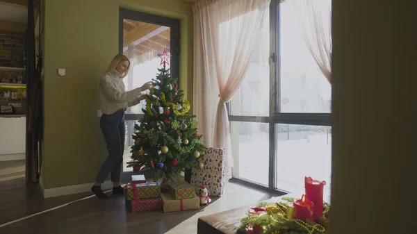 En vacker ung blond flicka i en tröja närmar sig julgranen och sätter på sig en julboll. En julgran vid fönstret i huset med en massa presenter under. — Stockfoto