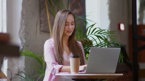 Nuori nainen istuu tietokoneen ääressä toimistossa ja juo kahvia. Liikenainen — kuvapankkivideo