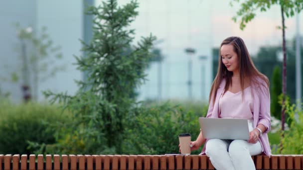 Una joven está sentada en una computadora en la calle y tomando café. Mujer de negocios — Vídeos de Stock