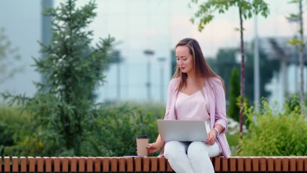 Una joven está sentada en una computadora en la calle y tomando café. Mujer de negocios — Vídeos de Stock