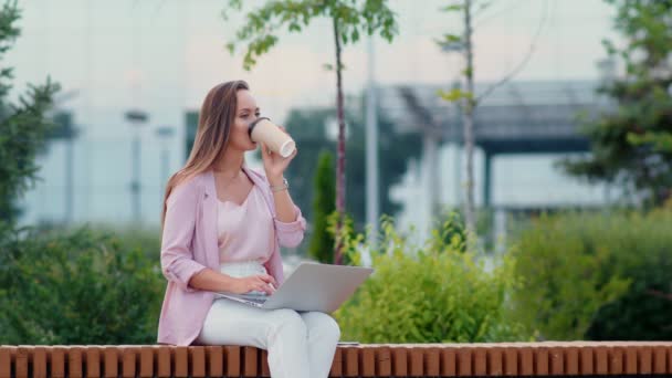 Nuori nainen istuu tietokoneen ääressä kadulla ja juo kahvia. Liikenainen — kuvapankkivideo